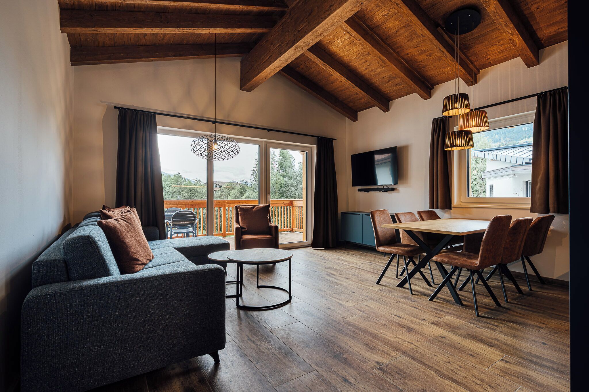 A spacious living area with a wooden ceiling, a sofa, a dining table with chairs, and large windows offering a view of the outdoors.