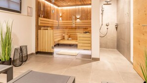 A sauna room with wooden walls and a glass door, a shower area with a wall-mounted showerhead, and a seating area with a bench and a small table. - Der Kehlbachwirt by Monteviva (Niedernsill)