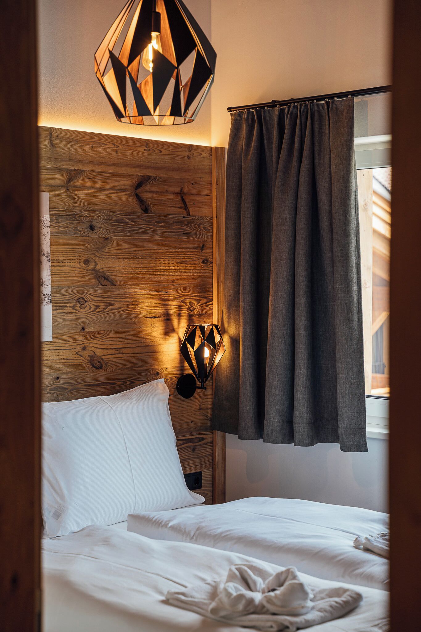Cama bem arrumada com lençóis brancos, cabeceira de madeira, luminária pendente geométrica e janela com cortinas cinzas.