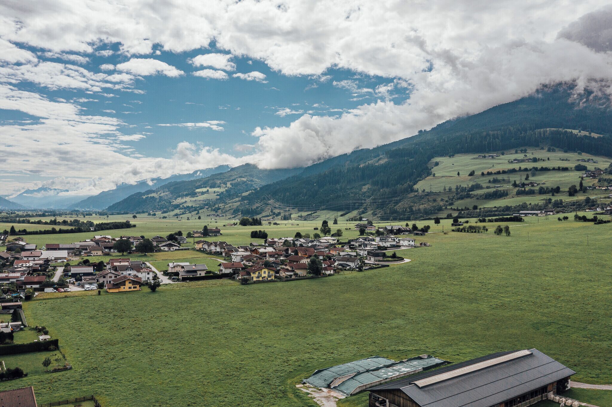 Un pueblo rural con casas y campos abiertos, rodeado de montañas y un cielo parcialmente nublado.