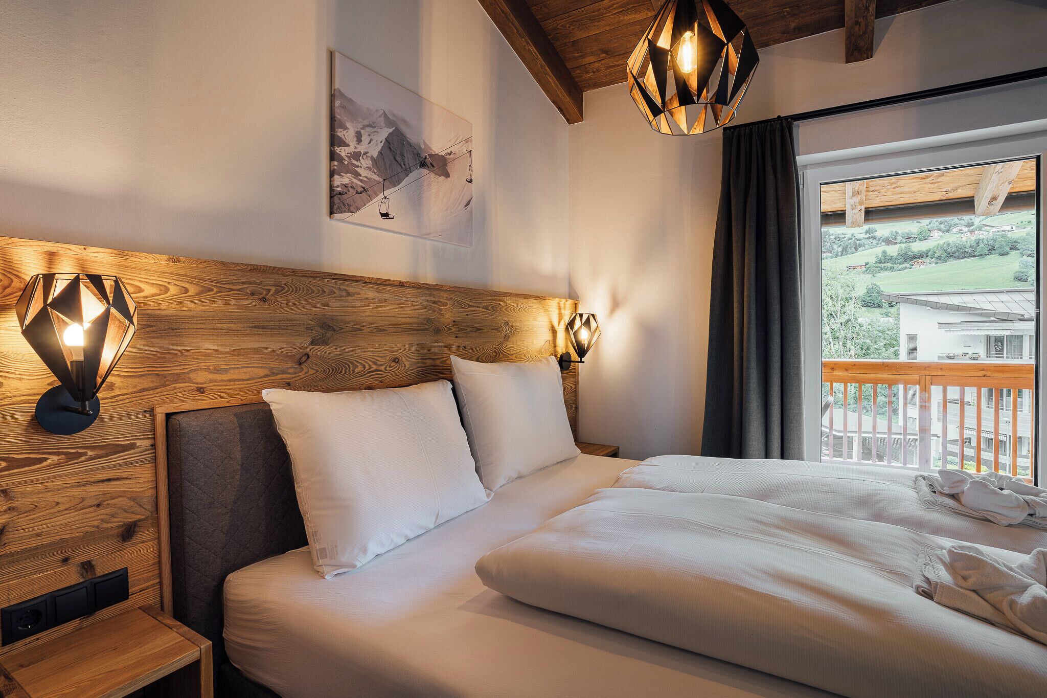 A hotel room with a wooden headboard, two bedside lamps, a large bed with white linens, and a view of a balcony with a mountain landscape.