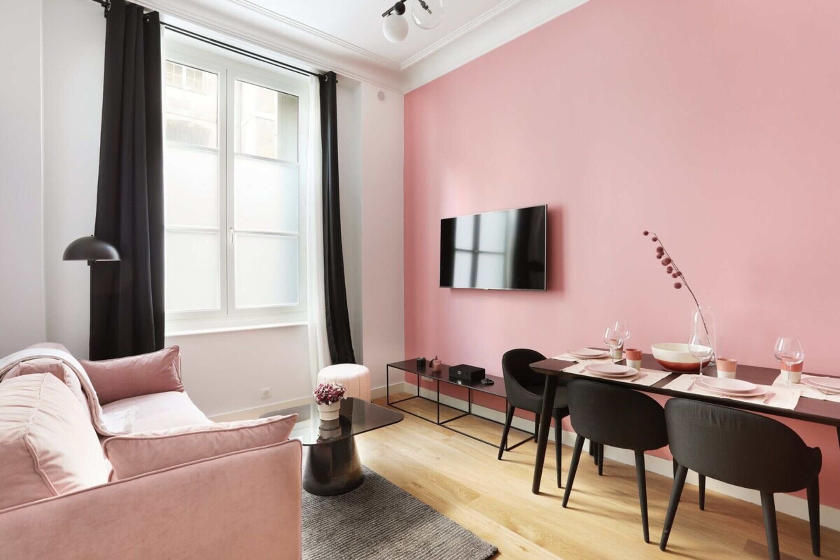 A modern living room with a pink wall, a flat-screen TV mounted above a dining table with chairs, a sofa, and a coffee table.