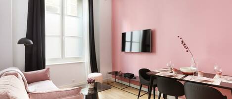 A modern living room with a pink wall, a flat-screen TV mounted above a dining table with chairs, a sofa, and a coffee table.