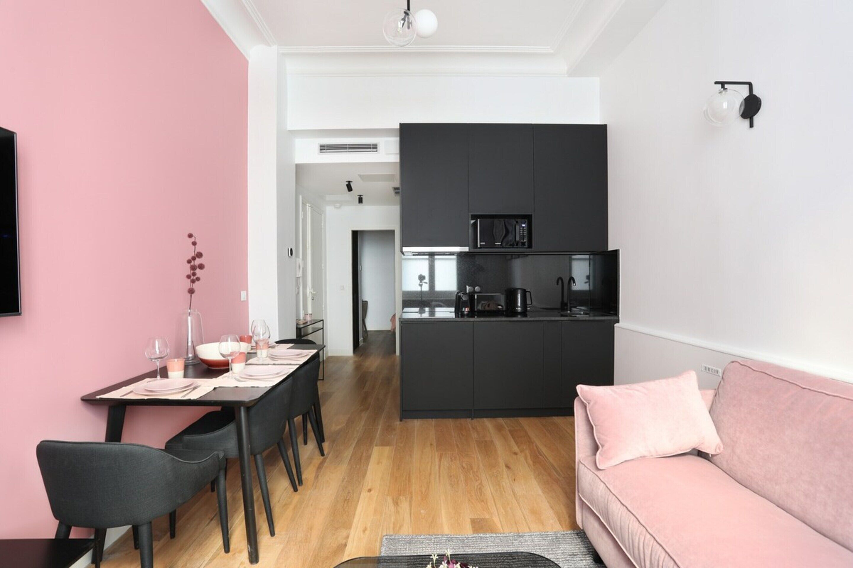 Een moderne keuken met zwarte kastjes en een magnetron, een eethoek met een roze muur, en een roze bank.