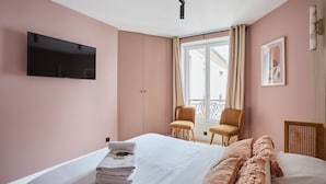 Una habitación de hotel con una cama, un televisor montado en la pared, dos sillas, un ventanal con cortinas y un cuadro enmarcado.