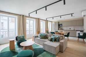 Un appartement moderne comprenant un espace de vie, un coin repas et une cuisine. Il est doté de grandes fenêtres, d’un ensemble de canapés et d’une table à manger.