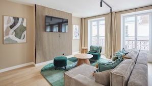 Una sala de estar moderna con un sofá, un reposapiés y una mesa de centro redonda. Un televisor de pantalla plana está montado en la pared, y hay ventanas grandes con cortinas.
