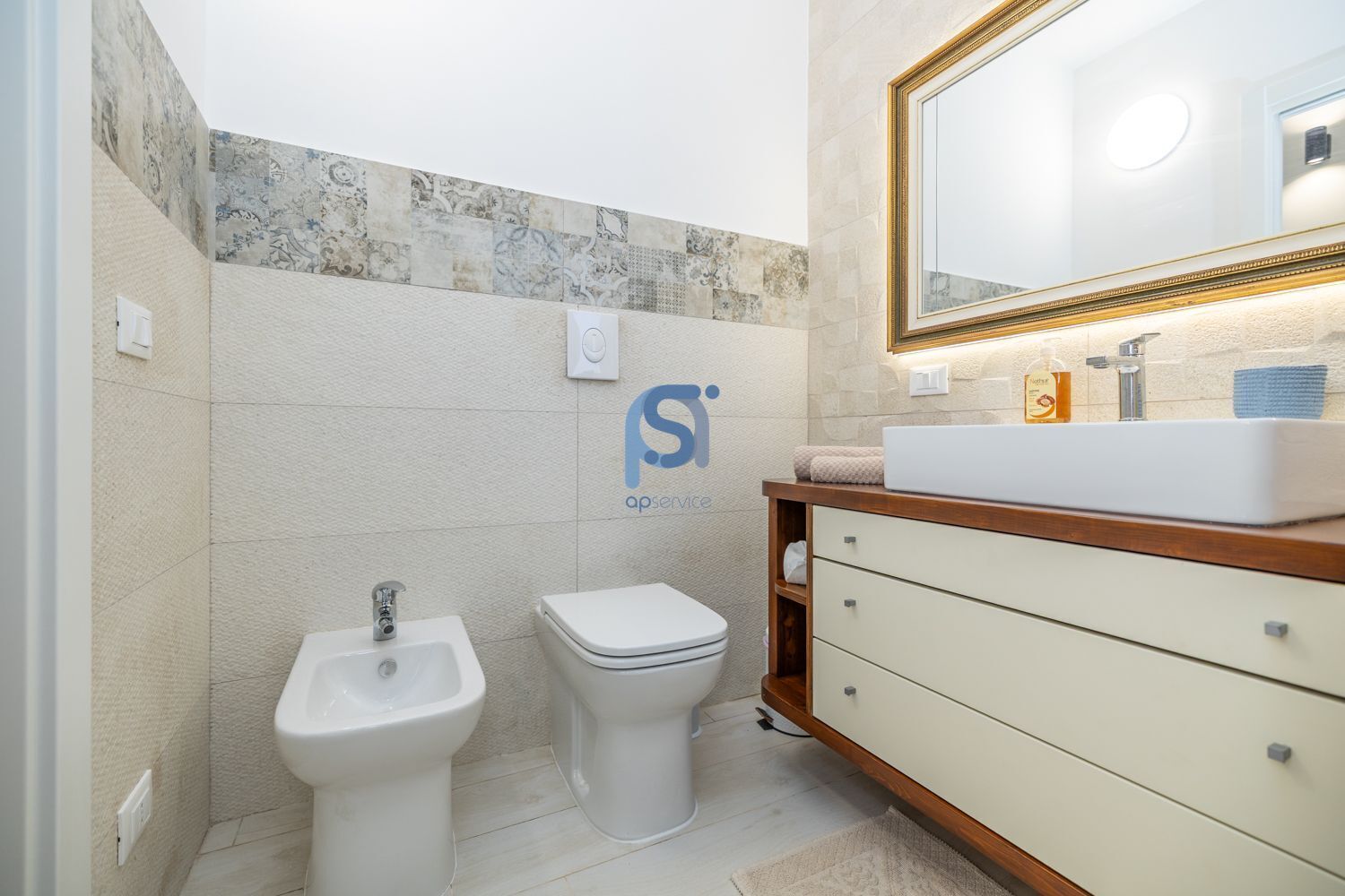 A bathroom with a sink, bidet, and toilet, a mirror with a gold frame, and a logo on the wall.