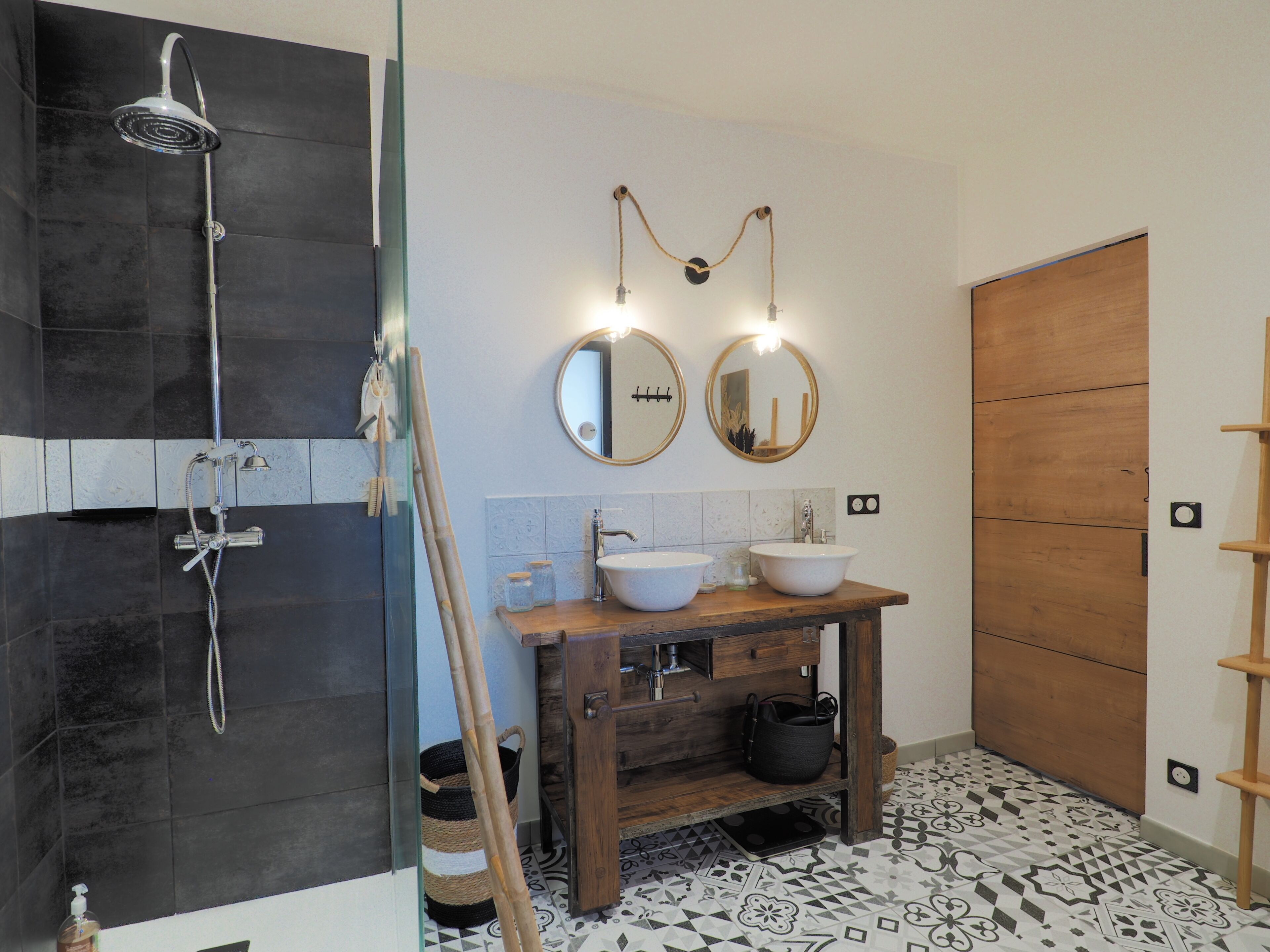 A modern bathroom with a glass shower enclosure, two sinks, wooden vanity, and patterned floor tiles.