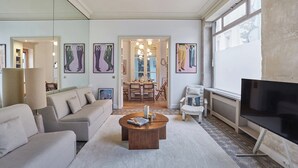 Uma sala de estar moderna com sofá, mesa de centro e televisão. Ao fundo, uma área de jantar com candelabro.