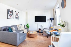 A modern living room with a grey sofa, a round wooden coffee table, a flat-screen TV mounted on the wall, a fireplace, and a dining area with a glass table and chairs. - Prime Fitzrovia with Balcony by Oxford Circus (London)