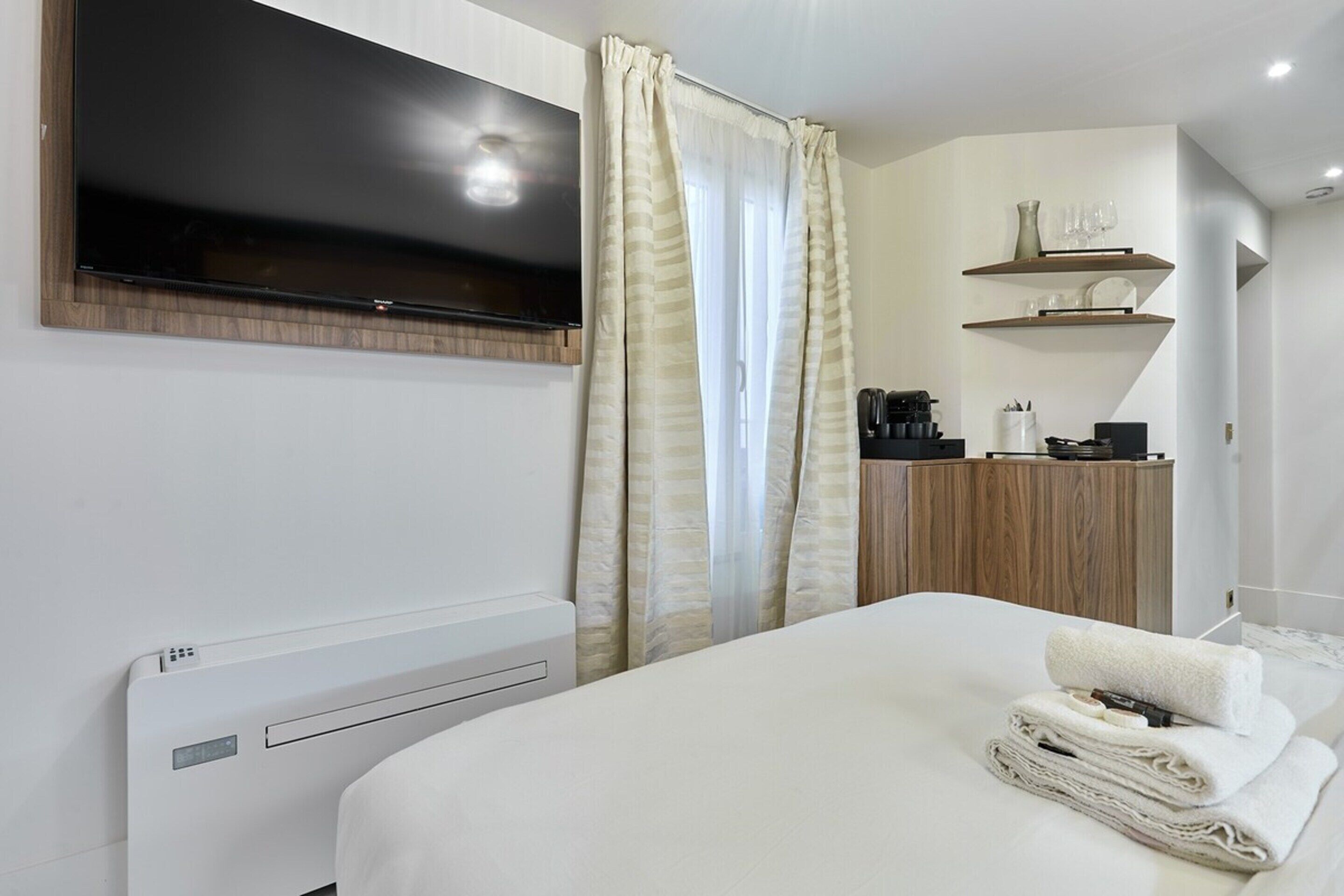 A modern hotel room with a bed, a flat-screen TV mounted above it, a coffee machine, and a shelf with glasses and a vase.