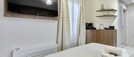A modern hotel room with a bed, a flat-screen TV mounted above it, a coffee machine, and a shelf with glasses and a vase.
