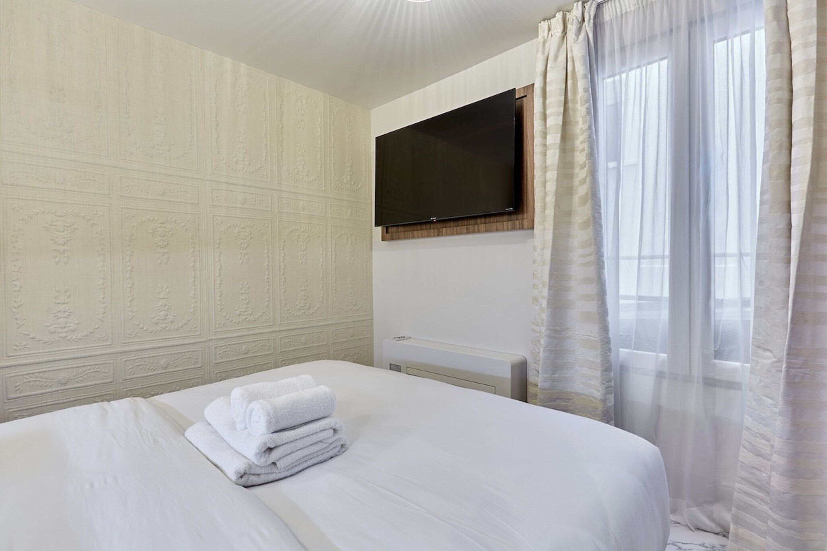 A hotel room with a bed, a flat-screen TV mounted on the wall, and a window with sheer curtains.