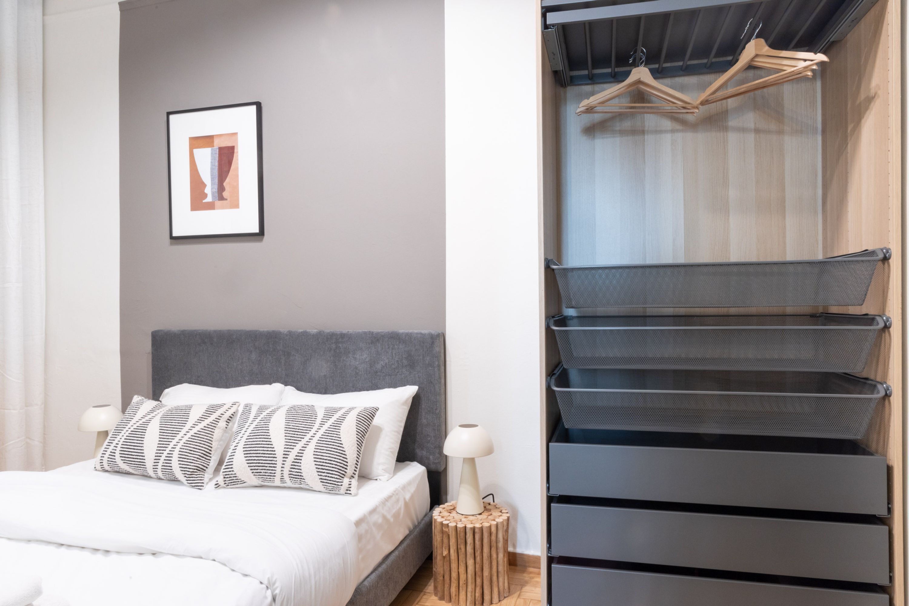 Un dormitorio moderno con cabecera gris, cama con ropa de cama blanca, un taburete de madera pequeño, una mesita de noche con lámpara, un cuadro enmarcado en la pared y un armario empotrado con espacio para colgar.