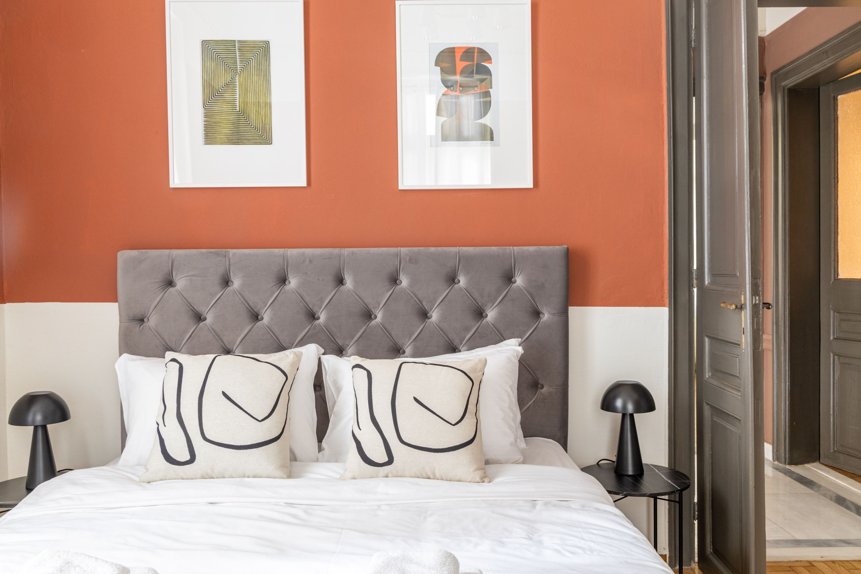 A neatly made bed with a tufted headboard, two framed artworks on the wall, and a door leading to another room.