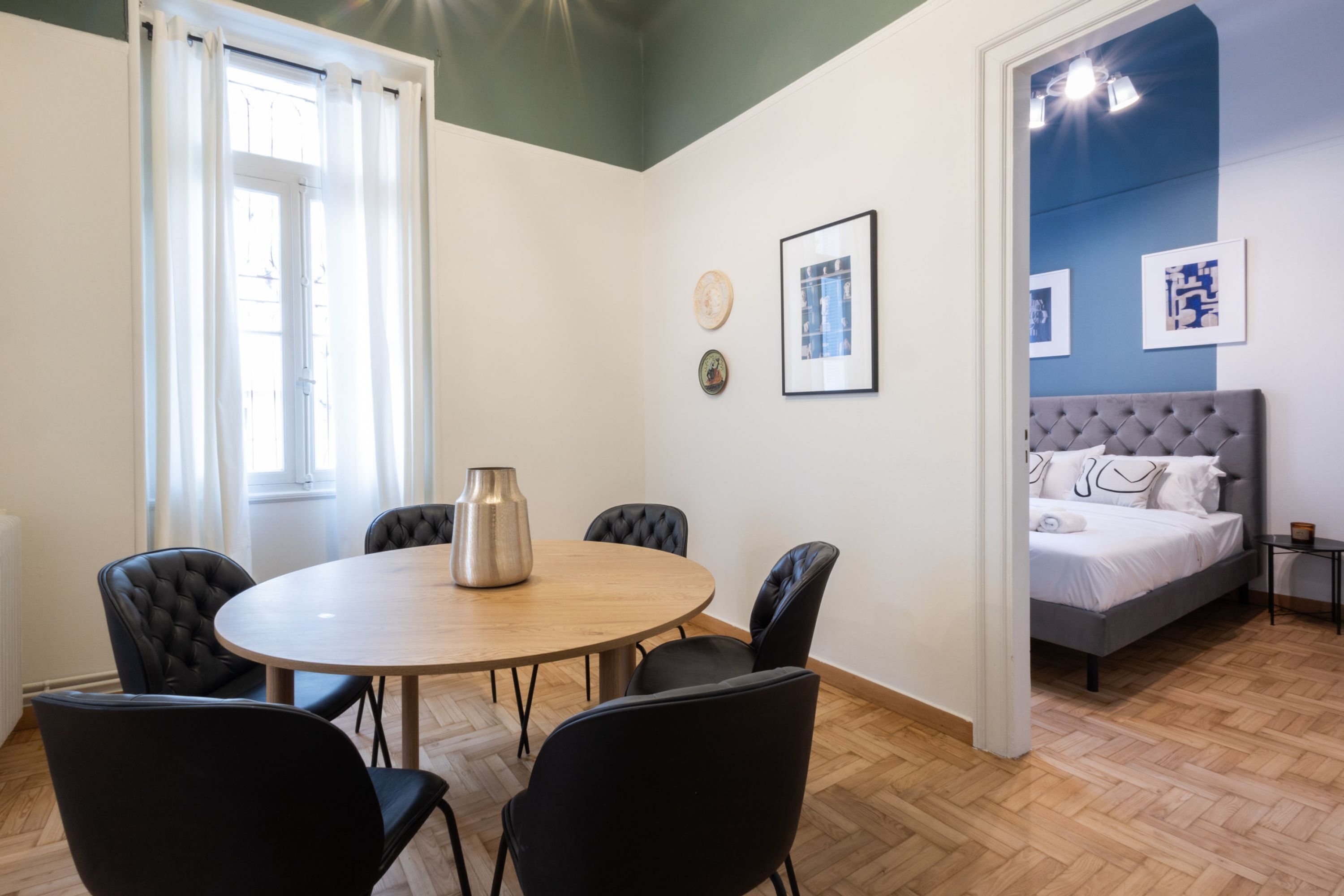 A room with a round table, six chairs, a bed, and artwork on the walls.