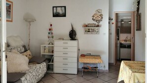 A living room with a sofa, a small table with a tablecloth, a cabinet, a floor lamp, and a wall-mounted picture. - 2 Bed apartment ground floor / Mont St Martin (CAP D AGDE)
