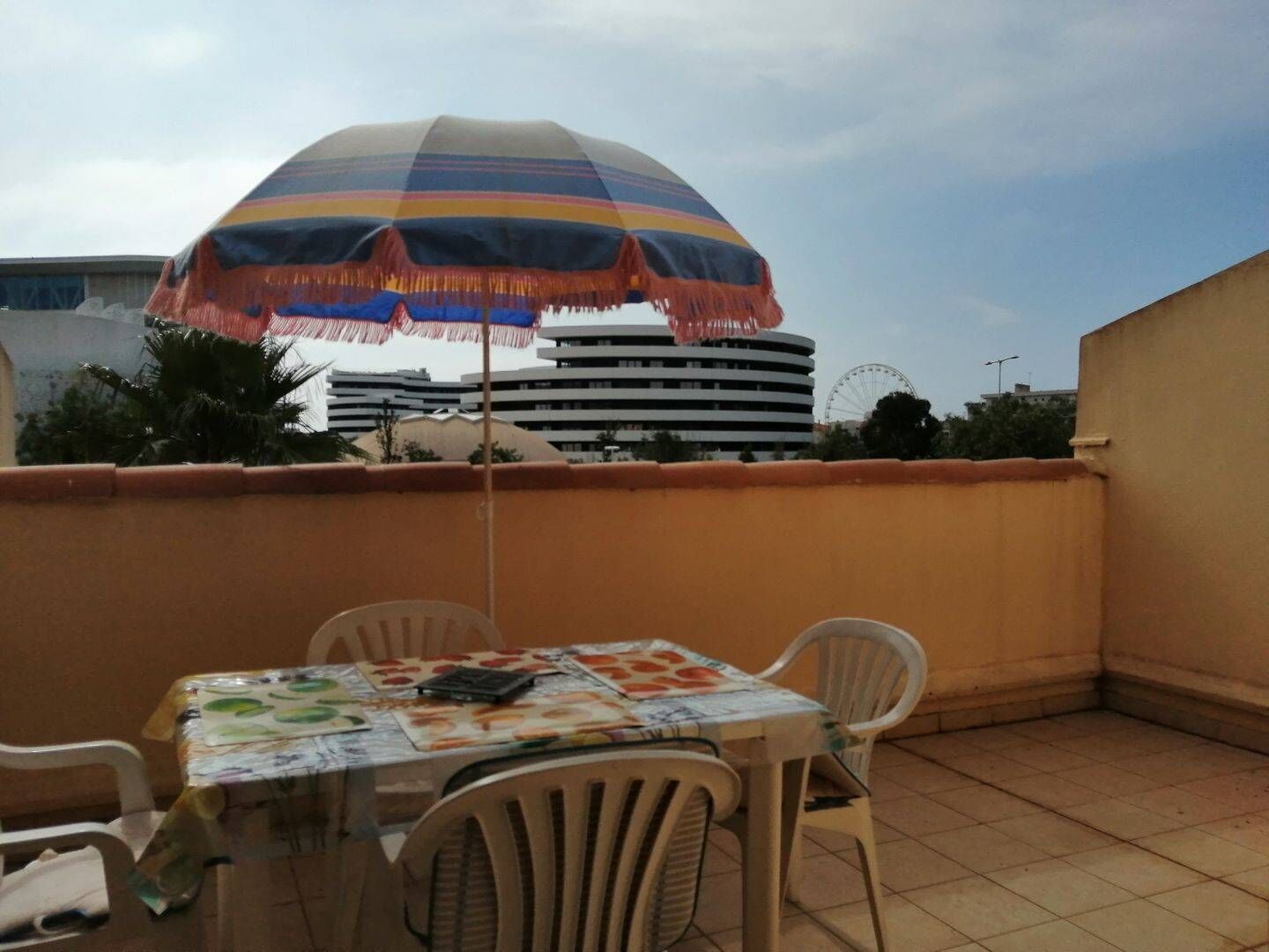 A small outdoor patio with a table and chairs, a colorful umbrella, and a view of a building with a Ferris wheel in the background.