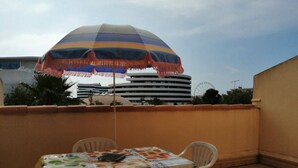 A small outdoor patio with a table and chairs, a colorful umbrella, and a view of a building with a Ferris wheel in the background.