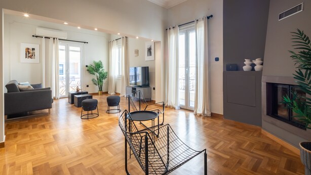 A spacious living room with a wooden floor, a grey sofa, a black metal chair, a small round table, and a fireplace. - Acropolis View 3-bdrm With Rooftop Garden Jacuzzi (Athens)
