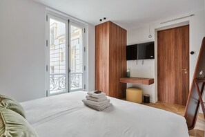 A bedroom with a bed, a wooden wardrobe, a TV, and a balcony with a view of buildings.