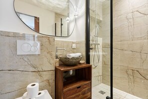 A modern bathroom with a marble shower wall, a wooden vanity with a bowl sink, and a large mirror.
