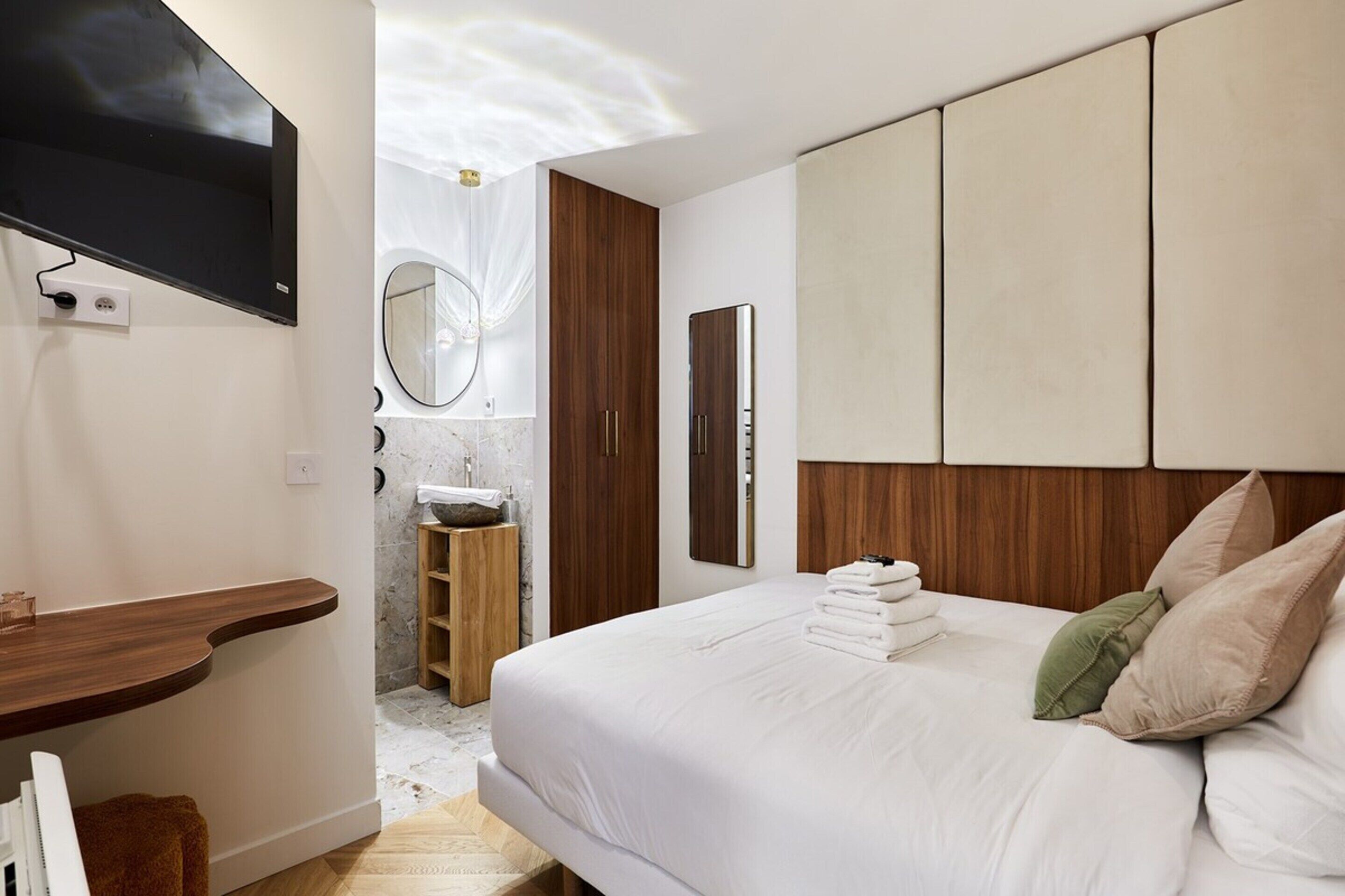 A hotel room with a large bed, a wooden headboard, a mirror, a TV mounted on the wall, and a wooden shelf.