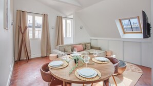 A dining area with a round table set for four, a sofa, a television, and a window with curtains. - Superb Flat Ave Montaigne With Eiffel Tower View (Paris)