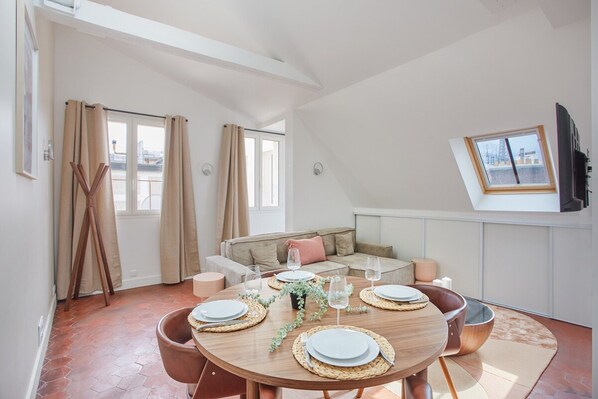 A dining area with a round table set for four, a sofa, a television, and a window with curtains.