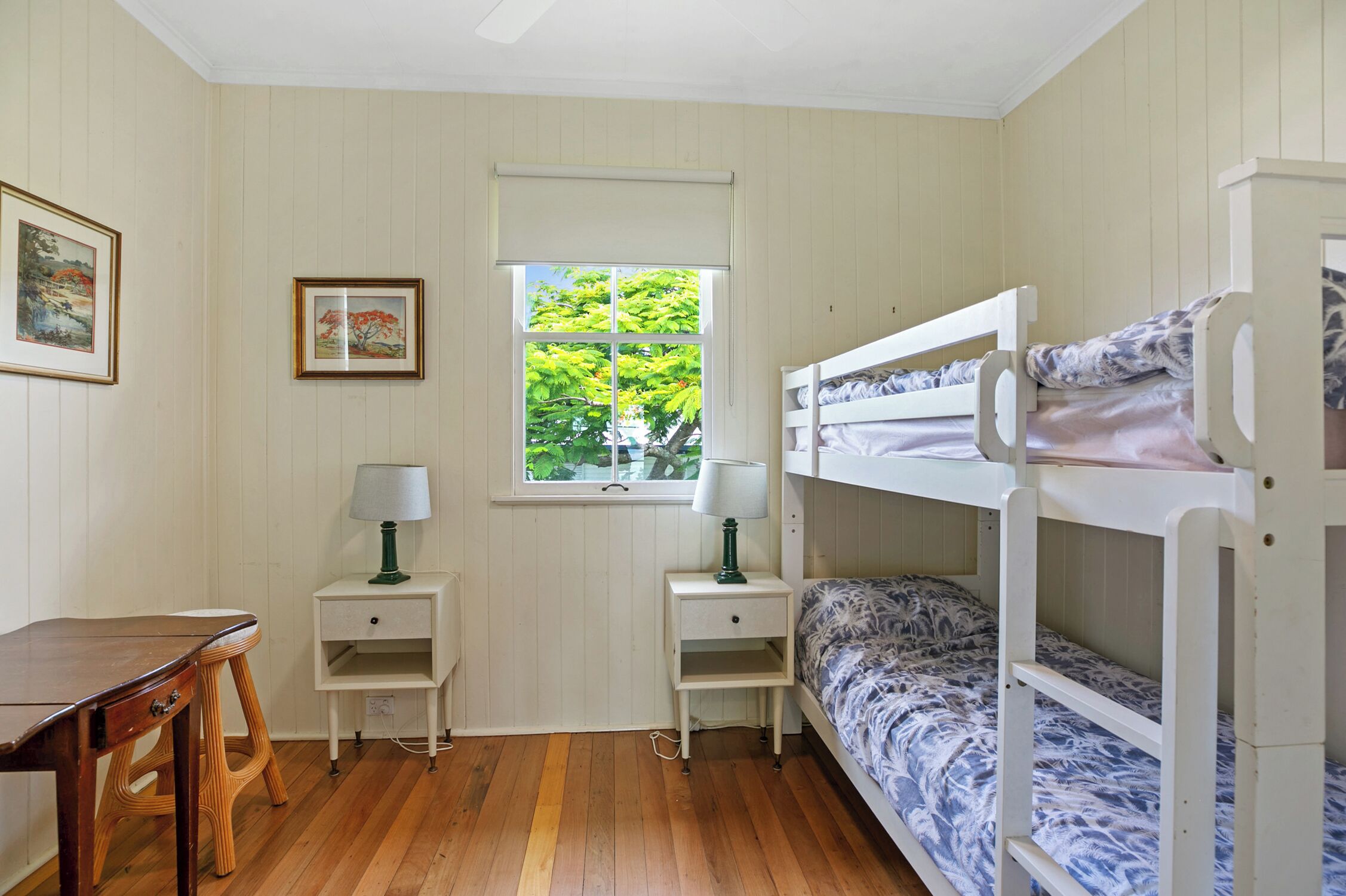 A room with a bunk bed, two bedside tables with lamps, a wooden desk, and framed pictures on the wall.