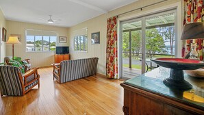 A spacious living room with wooden flooring, a ceiling fan, a television, a bench, and a glass table with a decorative item. - Welcome to Waterfront Views (Gympie)