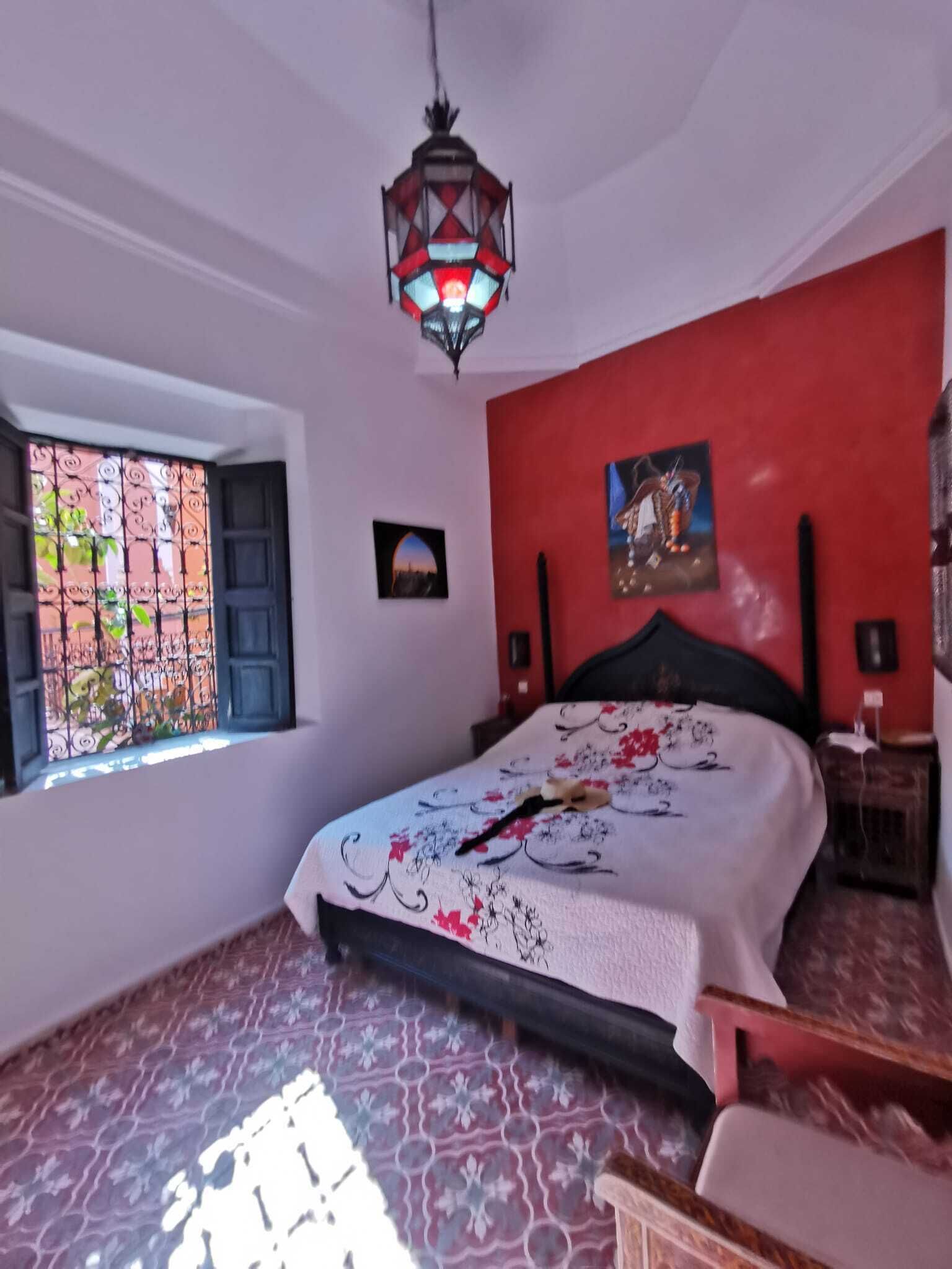 A bedroom with a bed, a window with a view, a red wall, and a decorative ceiling light.