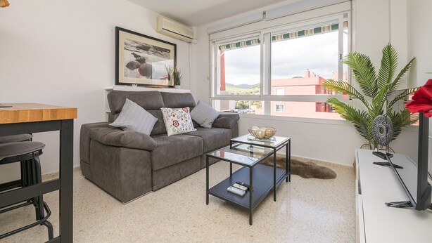 Una sala de estar moderna con un sofá gris, una mesa de centro de vidrio, una pequeña mesa de comedor y un ventanal con vistas a edificios.