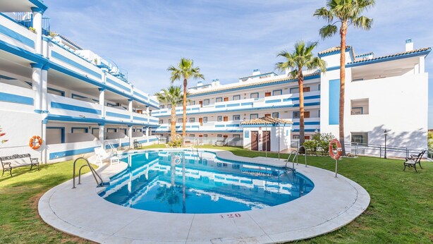 Une piscine entourée d’un bâtiment doté de nombreux balcons, de palmiers et d’une bouée de sauvetage.