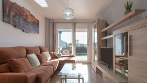 Un salon moderne avec un canapé marron, une table basse en verre, un téléviseur à écran plat, des étagères en bois et une grande fenêtre donnant sur un paysage verdoyant.
