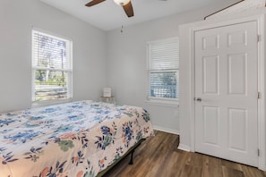 A bedroom with a floral bedspread, two windows with blinds, a ceiling fan, and a white door. - Charming Downtown High Springs Cottage (High Springs)
