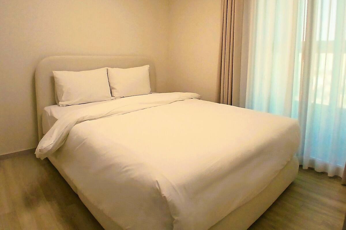 A neatly made bed with white linens in a room with wooden flooring and light-colored walls.