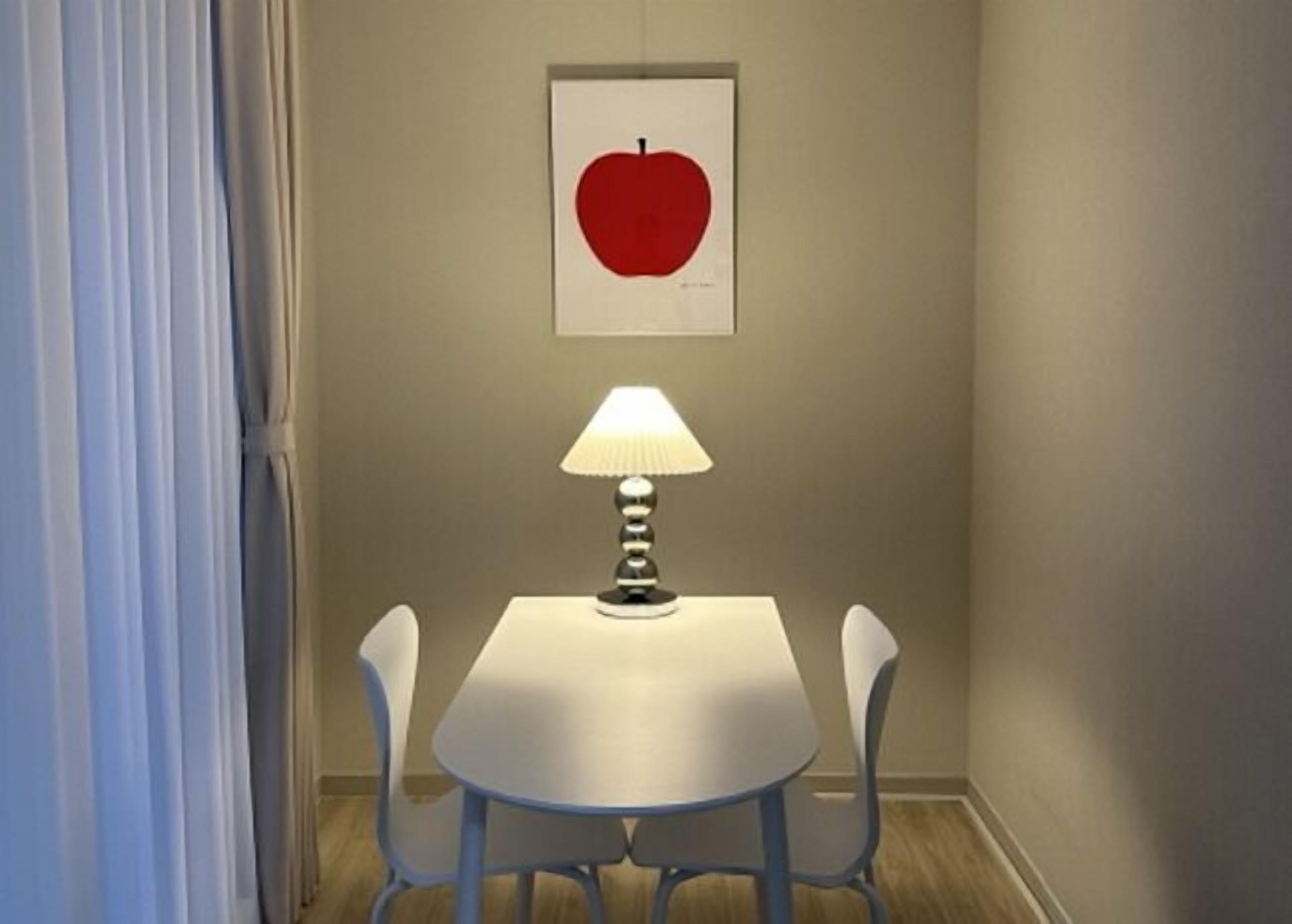 A dining area with a white table, two chairs, a table lamp, and a framed picture of an apple on the wall.