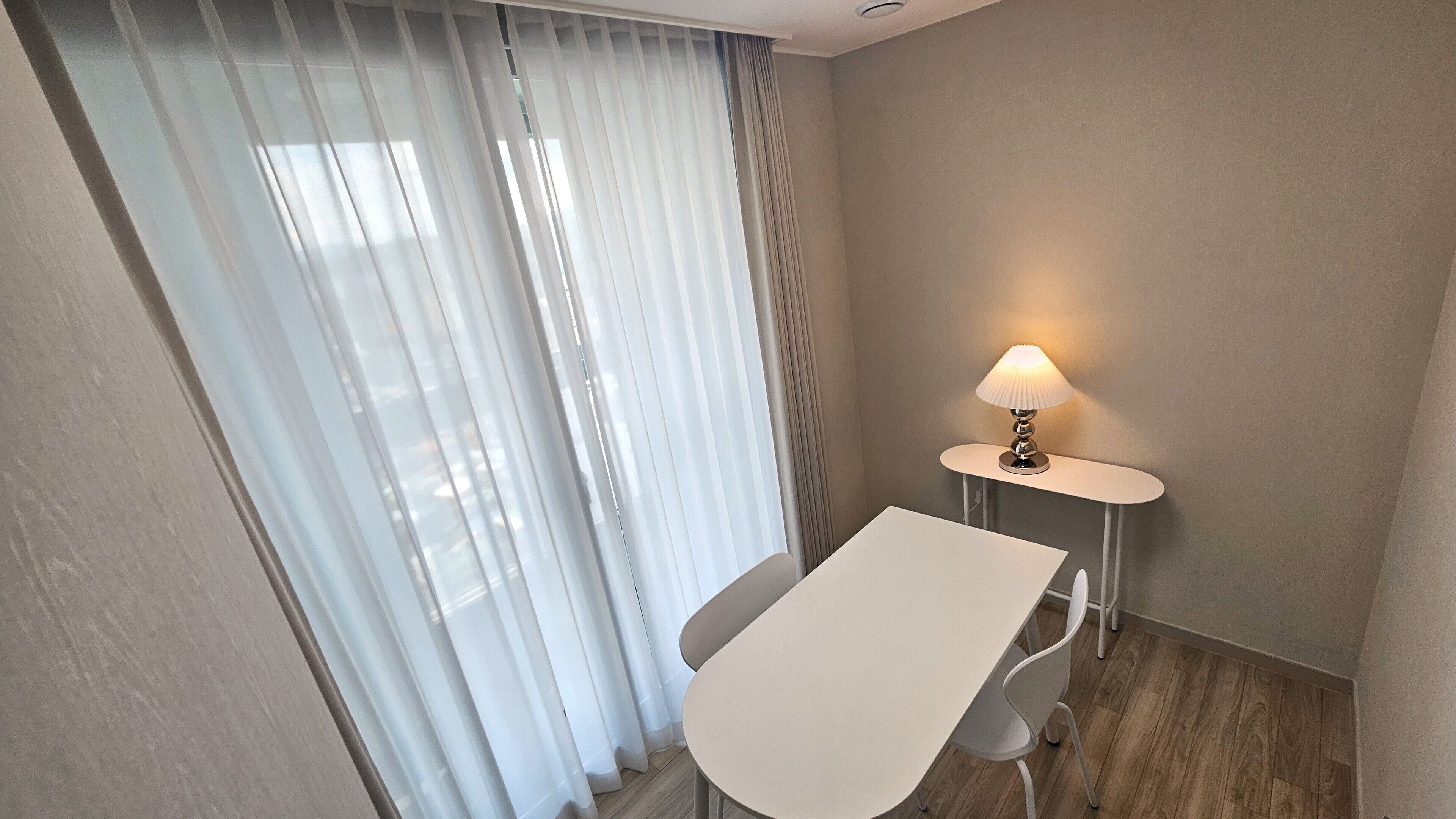 A small, modern room with a white table and chair, a lamp, and sheer curtains.
