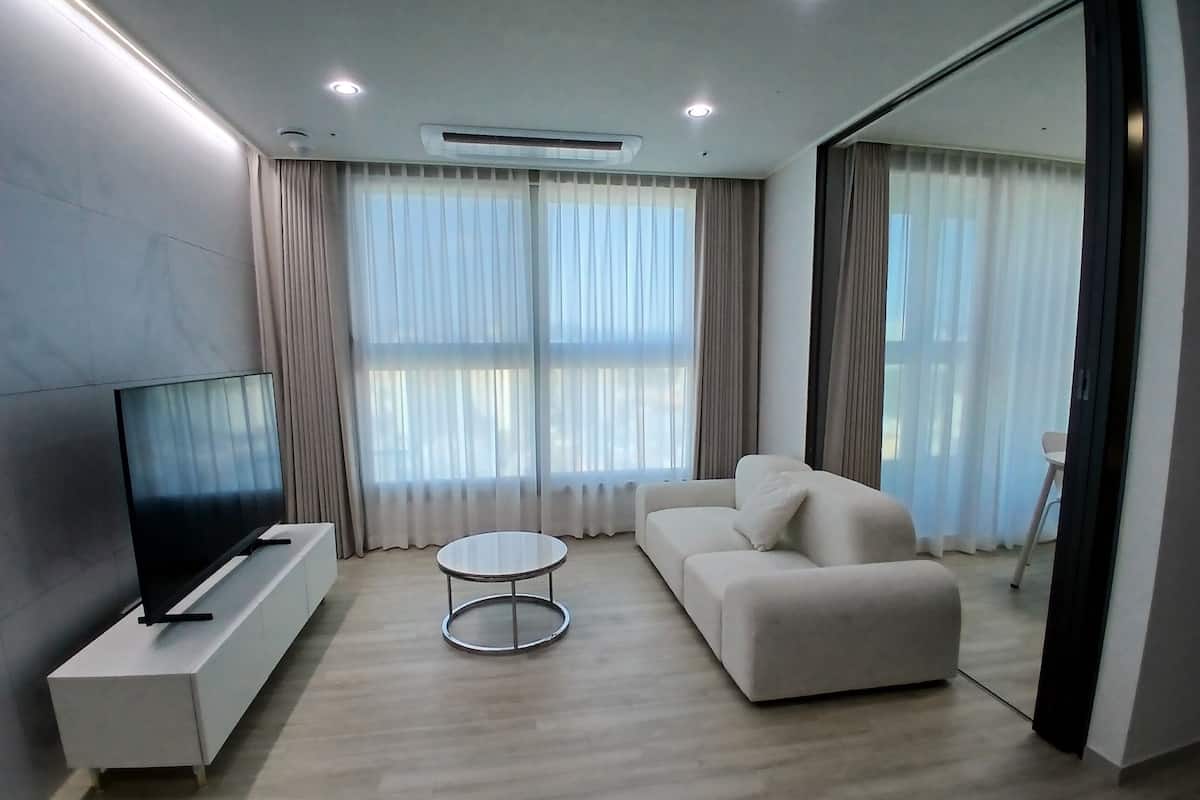 A modern living room with a flat-screen TV, a white sofa, a round coffee table, and large windows with sheer curtains.