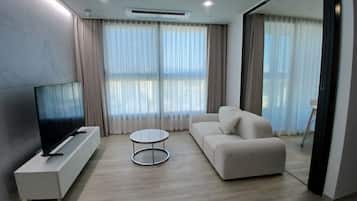 A modern living room with a flat-screen TV, a white sofa, a round coffee table, and large windows with sheer curtains.