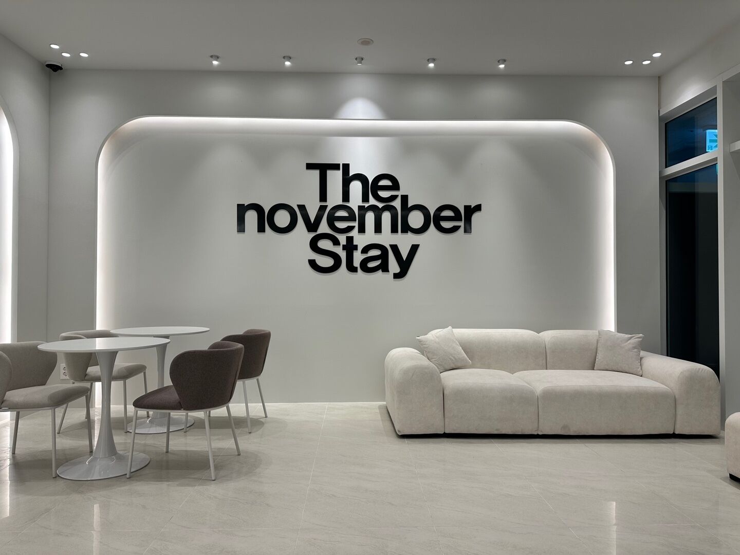 A modern interior with a large wall sign reading "The November Stay," a white sofa, and a dining area with round tables and chairs.