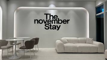 A modern interior with a large wall sign reading "The November Stay," a white sofa, and a dining area with round tables and chairs.