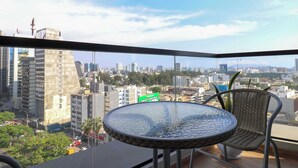 Un balcone con un tavolo rotondo e sedie, affaccia su un panorama urbano con edifici e cielo sereno.