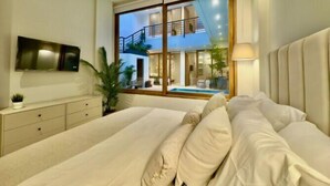 Ein Schlafzimmer mit einem großen Bett, einem an der Wand montierten Fernseher, einem Fenster mit Blick auf einen Pool und Sitzgelegenheiten im Freien sowie einem Nachttisch mit einer Pflanze.
