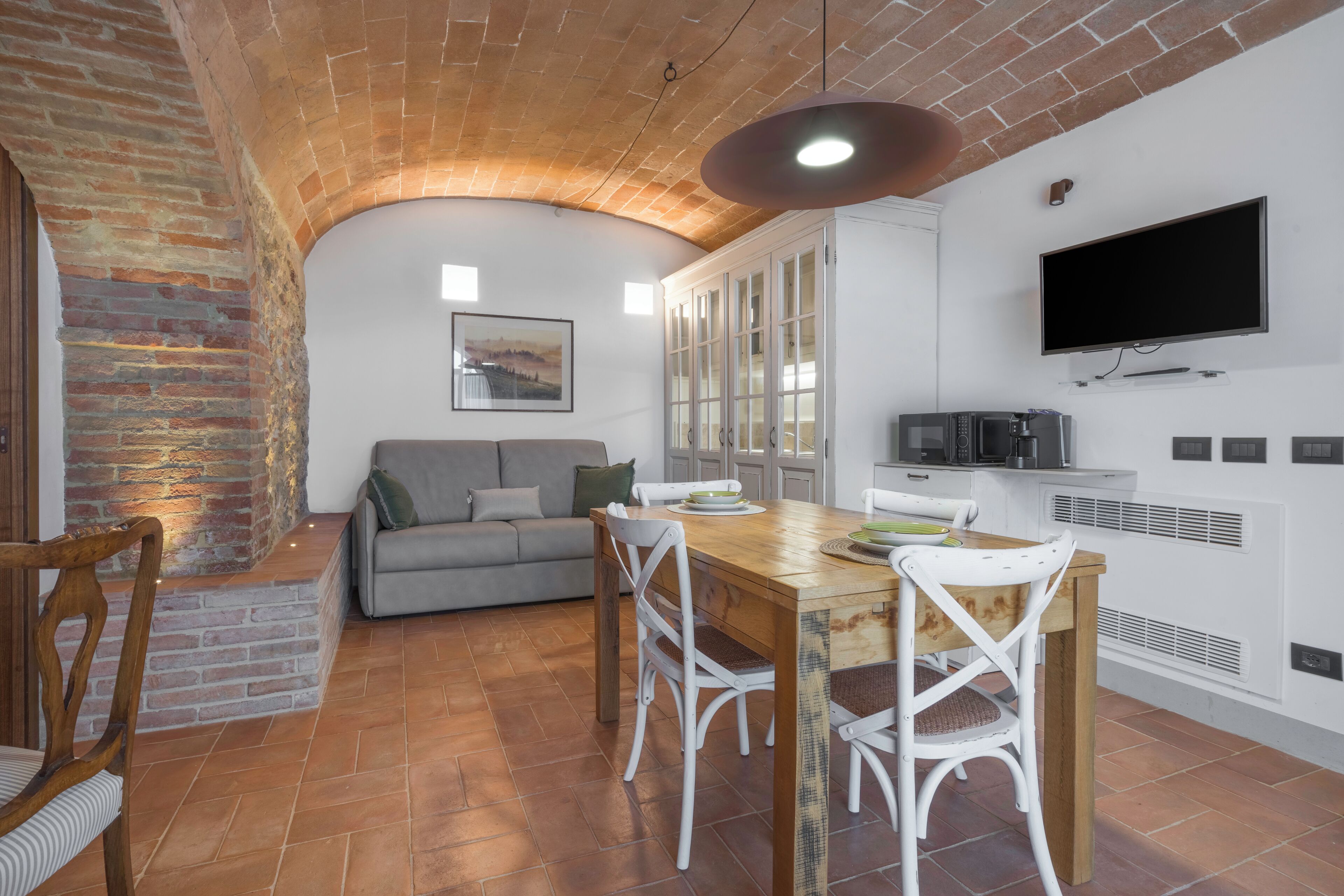 Una cucina compatta con zona pranzo, caratterizzata da un arco in mattoni, mobili bianchi e un televisore a schermo piatto.