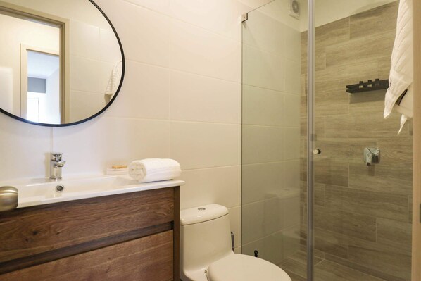 A bathroom with a wooden vanity, a white toilet, a glass shower enclosure, and a round mirror.