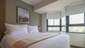 A neatly made bed with white linens and a beige throw blanket in a modern hotel room with a large window offering a city view. - w* | Great 2BR Apartment with Balcony in Miraflores (Lima)