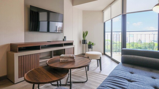 A modern living room with a sofa, a coffee table, a TV mounted on the wall, and a balcony with a view. - w* | Great 2BR Apartment with Balcony in Miraflores (Lima)